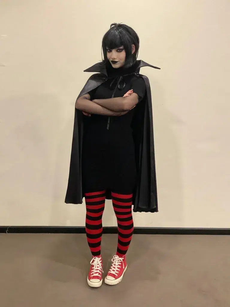 Petite woman wearing a black Halloween vampire character costume consisting of a fitted black ribbed short-sleeve knit mini dress layered under a full black satin cape with a dramatic pointed collar and ribbon tie at the neck, styled with red and black horizontally striped thigh-high stockings and red high-top canvas sneakers, wearing a short black bob wig with blunt bangs and dark lipstick, standing with arms crossed against a cream-colored wall.