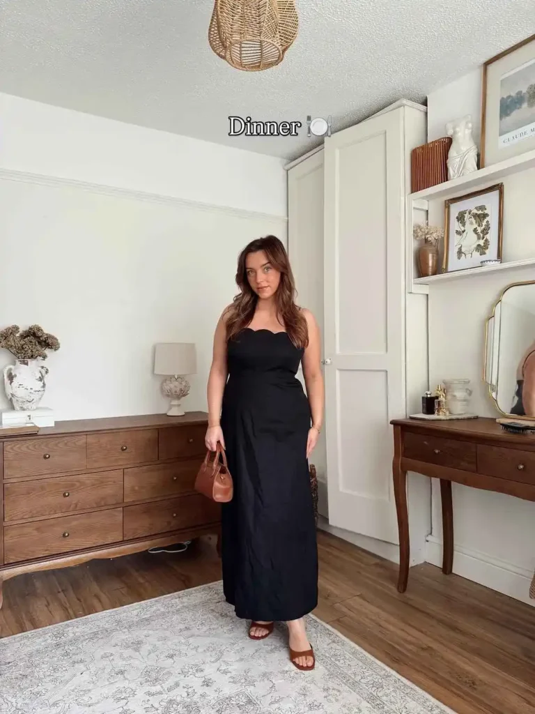 Petite woman in a black strapless maxi dress with a scalloped sweetheart neckline and fitted bodice, holding a small structured brown top-handle mini bag and wearing tan flat mule sandals, standing in a warmly decorated bedroom with dark wood furniture and neutral tones.