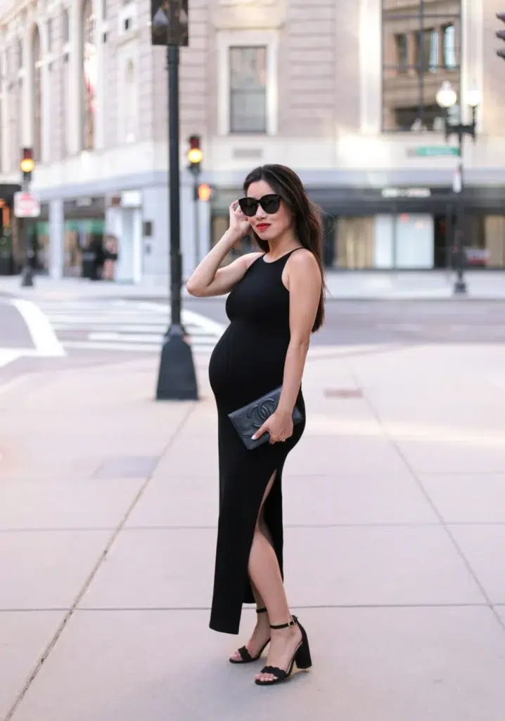 Petite pregnant Asian woman with long dark hair worn loose, wearing a sleek black sleeveless fitted maxi dress with a side slit at the lower hem and a racerback-style neckline, holding a small black quilted leather clutch with a round logo emblem in front of her bump, wearing black block heel open-toe ankle strap sandals, oversized black cat-eye sunglasses, and bright red lipstick, standing at a three-quarter side angle on a city corner sidewalk with tall stone and glass commercial buildings, traffic lights, and an open street visible in the background.