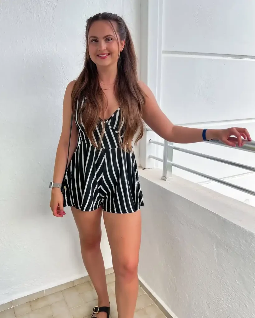 Petite woman in a black and white vertical stripe spaghetti strap romper with a wrap-style bodice and short flared hem, wearing flat black buckle sandals and delicate layered gold necklaces, standing on a bright sunlit balcony with white walls.