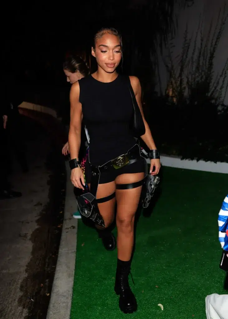 Woman with sleek pulled-back hair wearing an all-black action hero Halloween costume consisting of a fitted black sleeveless tank top, black micro shorts, a tactical utility belt with a large gold buckle, black leather thigh holster straps on both legs, black leather wrist cuffs, and tall black lace-up platform combat boots, walking outdoors at night on green artificial grass.
