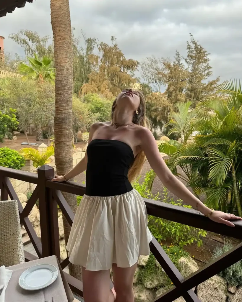Petite woman in a black structured strapless tube bodice and cream voluminous gathered bubble mini skirt, wearing layered gold necklaces and gold bracelets, standing on a wooden terrace balcony with tropical palm trees and lush greenery in the background.