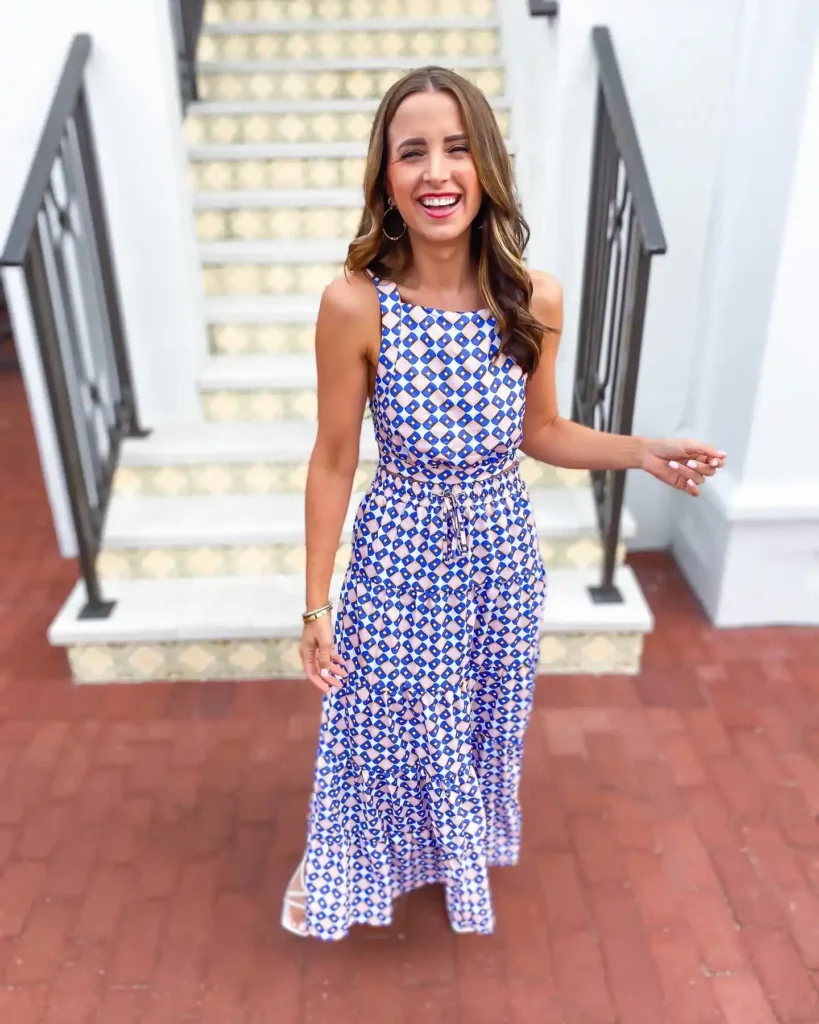 Petite woman in a sleeveless blue and nude geometric diamond-print two-piece maxi set with a soft drawstring waist and tiered ruffle hem, wearing white strappy heeled sandals and gold hoop earrings with a gold bangle bracelet, standing in front of decorative tiled stairs outdoors.