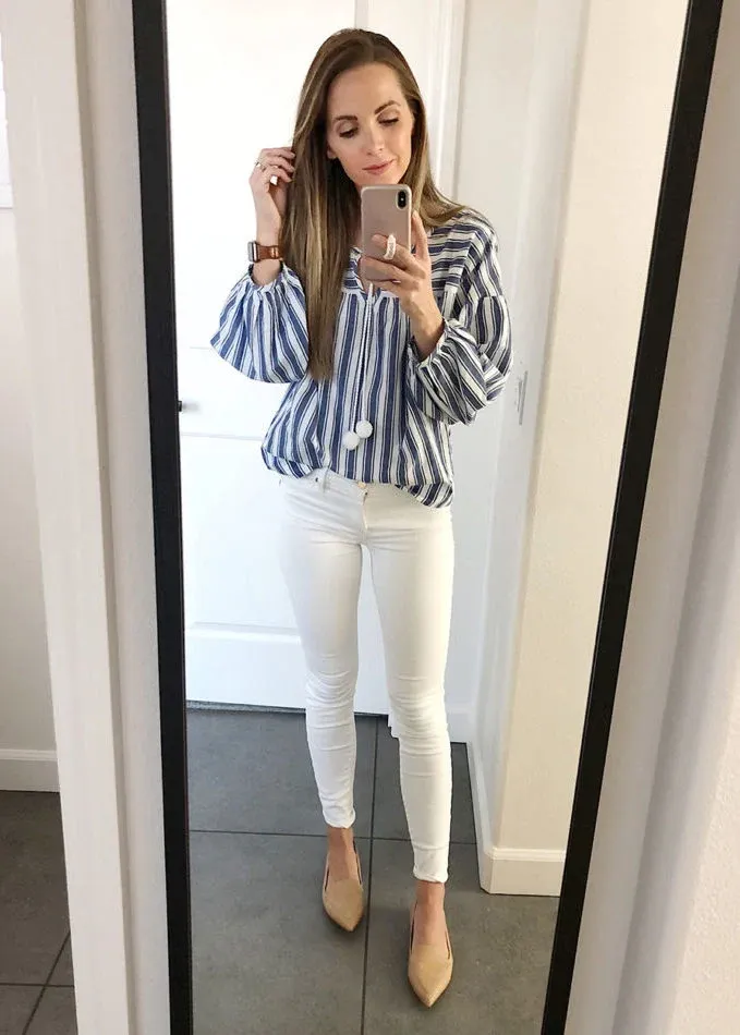 Petite woman with long straight brown hair taking a mirror selfie in a white-walled hallway with grey tile flooring, wearing white slim-fit ankle-crop skinny jeans with a clean hem, a bold blue and white wide vertical-stripe blouse with wide bell sleeves and white pom-pom tie detail at the center neckline, and nude camel pointed-toe low pumps, with a brown leather watch visible on her wrist.