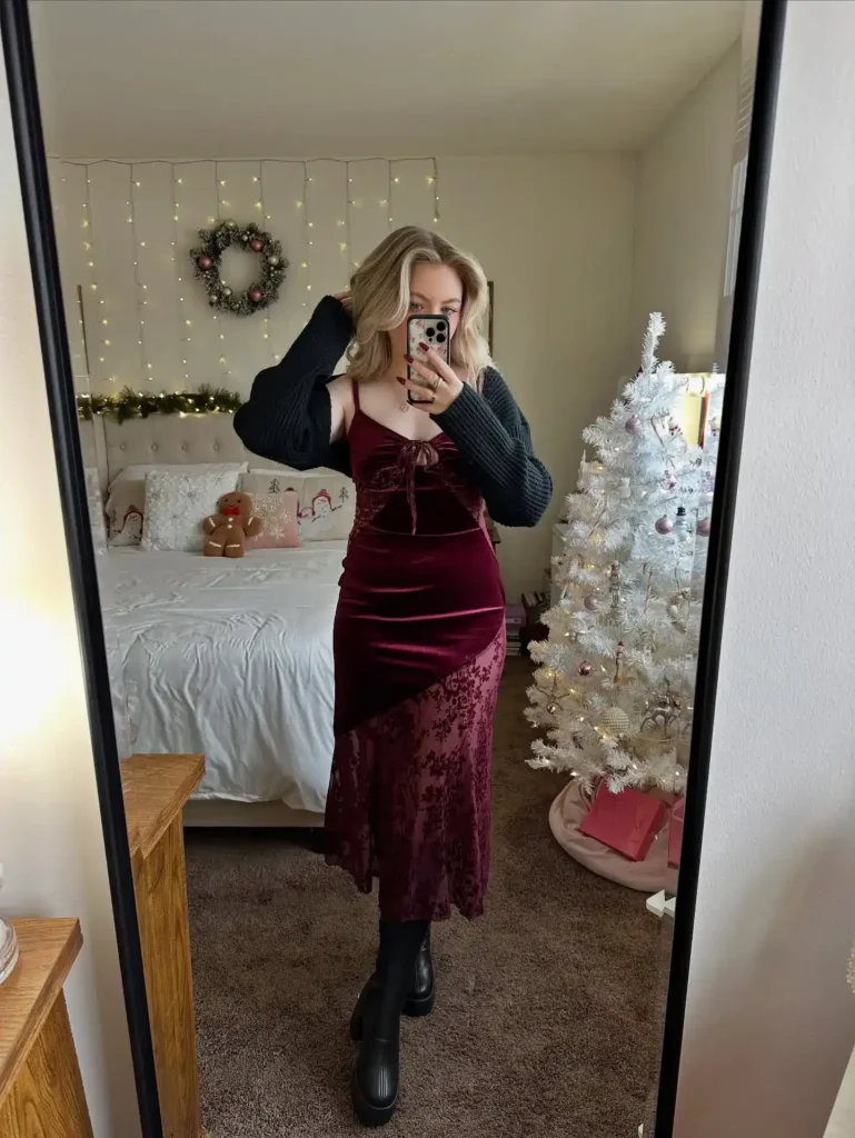 Woman taking a mirror selfie in a deep burgundy burnout velvet midi dress with a flocked floral pattern through the lower half, tie-front bow detail at the bust, and spaghetti straps, layered over a dark charcoal chunky cable-knit sweater with long sleeves visible at the arms, paired with black chunky lug-sole mid-calf boots, photographed in a cozy bedroom decorated with hanging fairy lights, a holiday wreath, and a white Christmas tree.