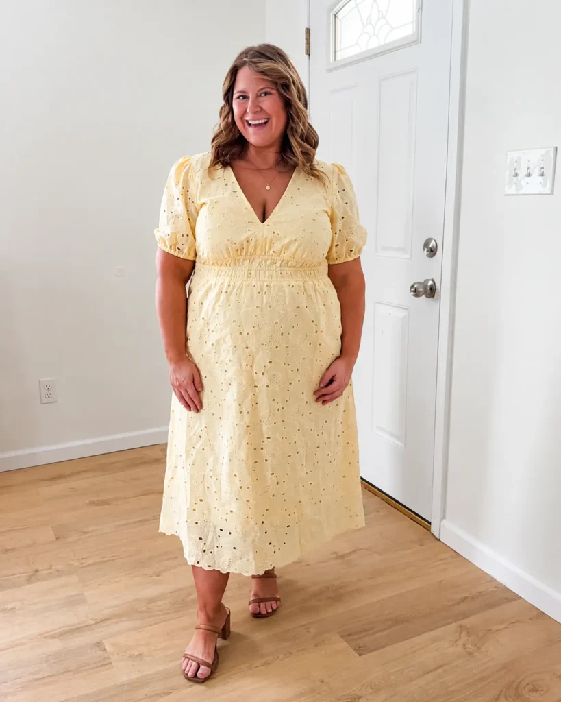 Petite plus-size woman with medium-length wavy honey-brown hair, smiling broadly with hands resting at her sides, wearing a soft butter yellow short-sleeve puff-sleeve midi dress made from broderie anglaise eyelet cotton fabric, with a V-neckline, elasticated gathered waistband defining the natural waist, a full A-line midi skirt, and a scalloped eyelet border at the hemline, paired with tan cognac block-heeled strappy sandals and a thin gold pendant necklace, standing on light wood flooring in a bright white-walled interior space near a white door.