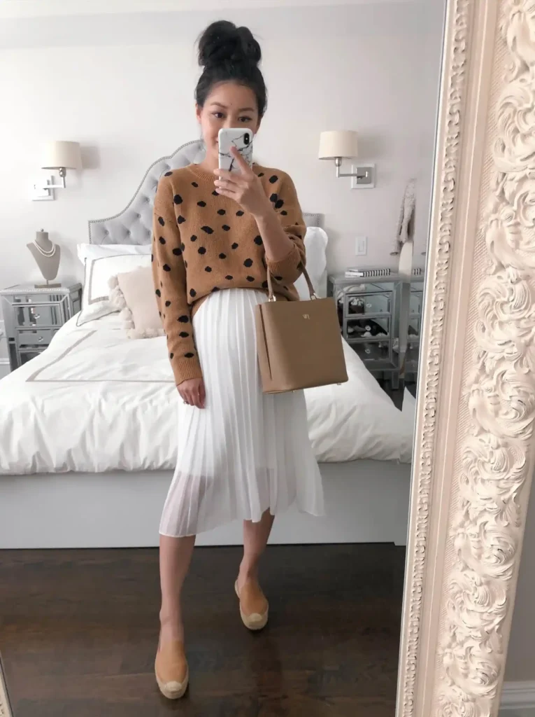 Petite pregnant Asian woman with dark hair in a high top bun, taking a bedroom mirror selfie wearing a camel tan knit sweater with large black polka dots oversized over a visible bump, paired with a white accordion pleated midi skirt, tan espadrille flatform wedge shoes, and a structured camel tan top-handle tote bag, standing in front of an ornate cream carved mirror frame with a grey tufted upholstered bed, mirrored nightstands, and wall sconce lamps visible in the background reflection.