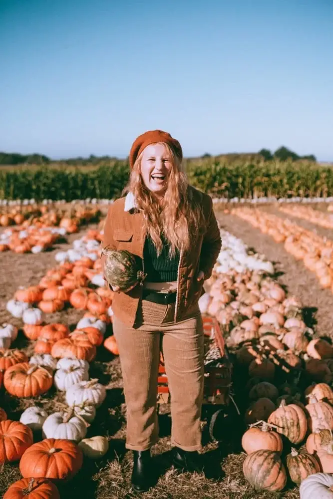 Petite woman with long wavy auburn hair wearing a rust beret tilted to one side, a caramel corduroy jacket with sherpa collar layered over a dark forest green ribbed knit top, matching caramel corduroy wide-leg cropped pants, a black belt bag cinched at the natural waist, and black ankle boots, laughing freely while holding a small green gourd in a wide sunny pumpkin patch with rows of orange, white, and green pumpkins and a cornfield in the background.