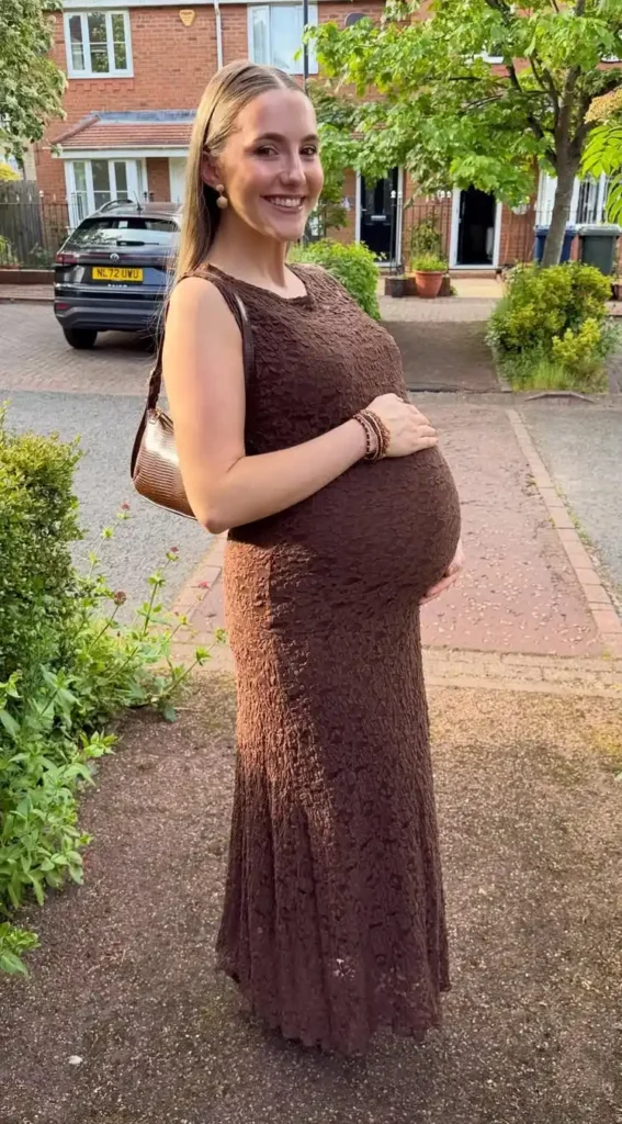 Pregnant woman in late third trimester with long straight blonde hair slicked back low, wearing a deep chocolate brown sleeveless fitted lace maxi dress with a simple scoop neckline that stretches smoothly over a very full bump, carrying a small brown textured shoulder bag over her left shoulder, wearing stacked warm-tone bracelets on her right wrist and small cream button stud earrings, standing on a residential street driveway with red brick UK terraced houses, parked cars, and leafy trees in the background.