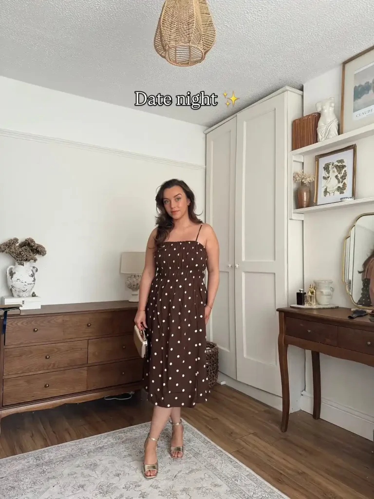 Petite woman in a chocolate brown and white medium-scale polka dot spaghetti strap smocked bodice midi dress with a full gathered skirt, wearing gold strappy heeled sandals and holding a small rectangular gold metallic clutch, standing in a warmly decorated bedroom with dark wood furniture and neutral decor.