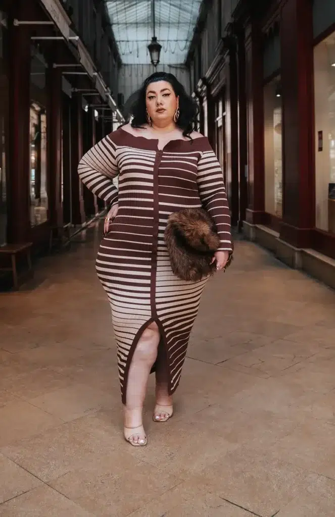 Petite plus-size woman with short dark hair and oversized gold hoop earrings wearing a chocolate brown and cream horizontal-striped ribbed knit long-sleeve bodycon midi dress with a wide bardot neckline, center-front seam detail, and front hem slit, holding a dark brown faux fur round clutch, styled with nude clear-strap heeled sandals, standing inside a covered glass-ceiling European shopping arcade with dark wood shopfronts lining both sides.