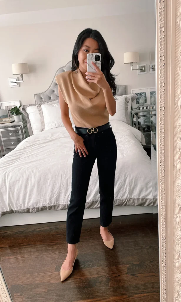 Petite woman in black slim-fit ankle-length jeans with a sleeveless camel cowl-neck knit top fully tucked in at the natural waist, secured with a narrow black leather belt with a gold horsebit-style buckle, paired with nude pointed-toe flats, taking a mirror selfie in a bedroom with a white upholstered headboard and an ornate gold-framed mirror.