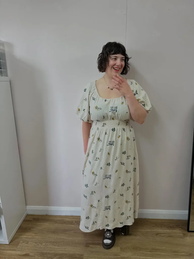 Petite plus-size woman with short dark wavy hair and blunt fringe, laughing with one hand raised near her face, wearing a cream off-white linen-look midi dress with a wide scoop neckline, short puff sleeves with a slightly flared cuff, a smocked waistband, and a full relaxed A-line midi skirt printed with small scattered botanical motifs including lavender sprigs, chamomile-style flowers, and wildflowers in sage green, mustard yellow, and teal, paired with chunky black platform shoes with a floral embossed Mary Jane strap detail, a delicate gold charm necklace, and a red lip, standing indoors against a light grey-pink wall on light wood flooring.
