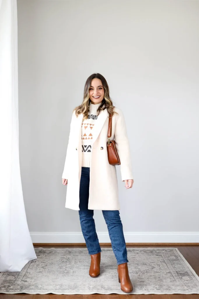 Petite woman with wavy highlighted brown hair standing against a neutral grey wall on a vintage-style area rug, wearing a cream longline double-breasted wool-blend coat worn open, a Fair Isle geometric-patterned sweater in cream with rust and dark tones visible at the chest, dark straight-leg jeans, and cognac block-heel ankle boots, carrying a cognac structured saddle crossbody bag with a gold-tone buckle detail, smiling with arms relaxed at her sides.