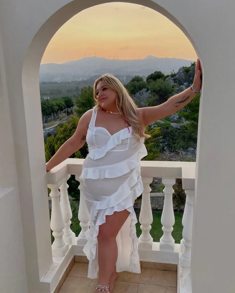 Petite plus-size woman with long loose blonde wavy hair, smiling and leaning with arms spread wide against the inner edge of a white arched balcony opening, wearing a crisp all-white sleeveless high-low dress with a structured sweetheart bustier bodice, one thick shoulder strap, and cascading horizontal tiered ruffle panels that grow fuller toward the asymmetric hem, styled with a layered shell and pearl choker necklace, thin stacked ankle bracelets, and strappy silver heeled sandals, with a warm golden sunset sky, green Mediterranean hillside, and white balustrade railing visible behind her.