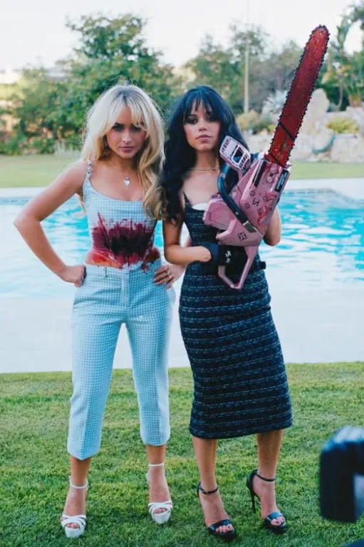 Petite woman with long dark wavy hair and blunt bangs wearing a fitted dark navy strapless tweed midi dress with horizontal ribbed textured banding, paired with black strappy stiletto heeled sandals, holding a decorative pink Halloween prop chainsaw, posing outdoors by a pool alongside another woman in a light blue gingham Halloween costume set.