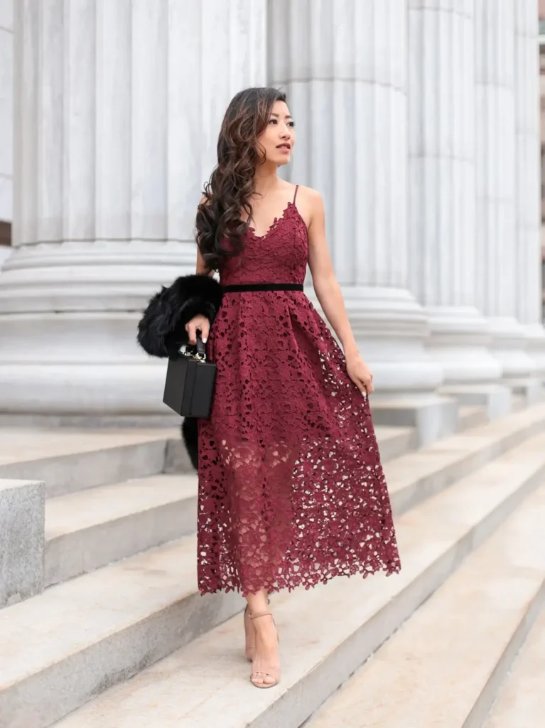 Petite woman in a deep wine burgundy guipure lace midi dress with spaghetti straps, a V-neckline with scalloped lace edging, a thin black velvet ribbon waist sash, and a full A-line lace skirt falling to mid-calf with a scalloped hem, wearing nude blush strappy block-heel sandals, carrying a small structured black box bag with gold hardware, with a black faux fur stole draped over one arm and long dark wavy hair, walking on wide stone steps in front of tall classical marble columns.