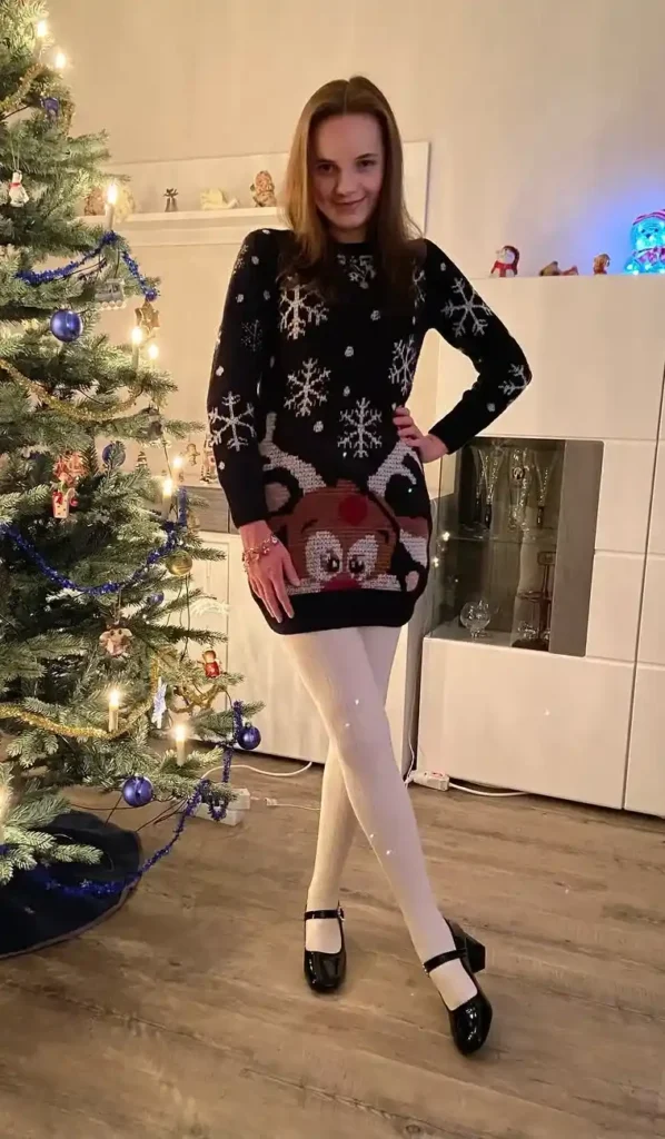 Petite woman in a dark navy oversized Christmas knit sweater dress with a Rudolph red-nosed reindeer and snowflake graphic print, wearing white opaque tights and black patent leather Mary Jane shoes with an ankle strap, a gold charm bracelet on her wrist, standing in front of a Christmas tree decorated with blue baubles and gold tinsel garland in a warmly lit living room.