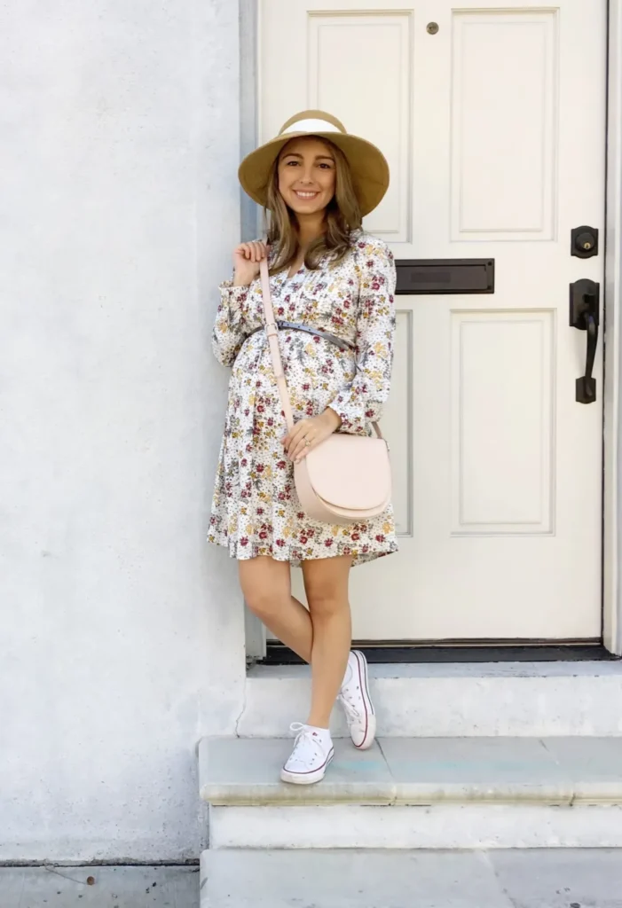 Pregnant woman with long wavy light brown hair wearing a wide-brim tan straw hat, dressed in a white long-sleeve floral mini dress with a small print in burgundy, mustard yellow, and olive green tones with a thin tie detail below the bust, carrying a small round blush pink crossbody saddle bag, wearing white high-top canvas sneakers, leaning casually against the doorframe of a cream-painted front door on stone entry steps.