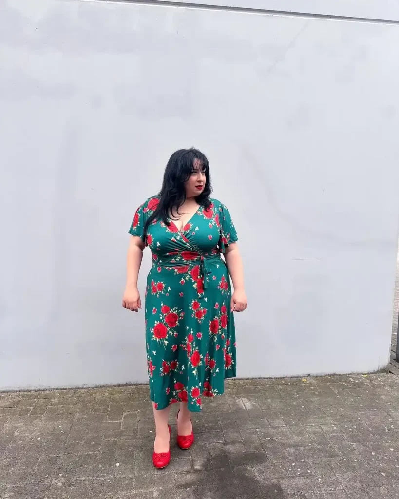 Petite plus-size woman with short dark wavy hair and blunt fringe, wearing a forest teal green wrap midi dress printed with large red roses, small greenery, and scattered white butterflies, with short flutter sleeves, a deep V wrap neckline, a self-tie waist detail, and a soft A-line midi skirt landing below the knee, styled with matching red pointed-toe pumps with a ruched toe cap and a bold red lip, standing in front of a plain grey wall on cobblestone pavement.