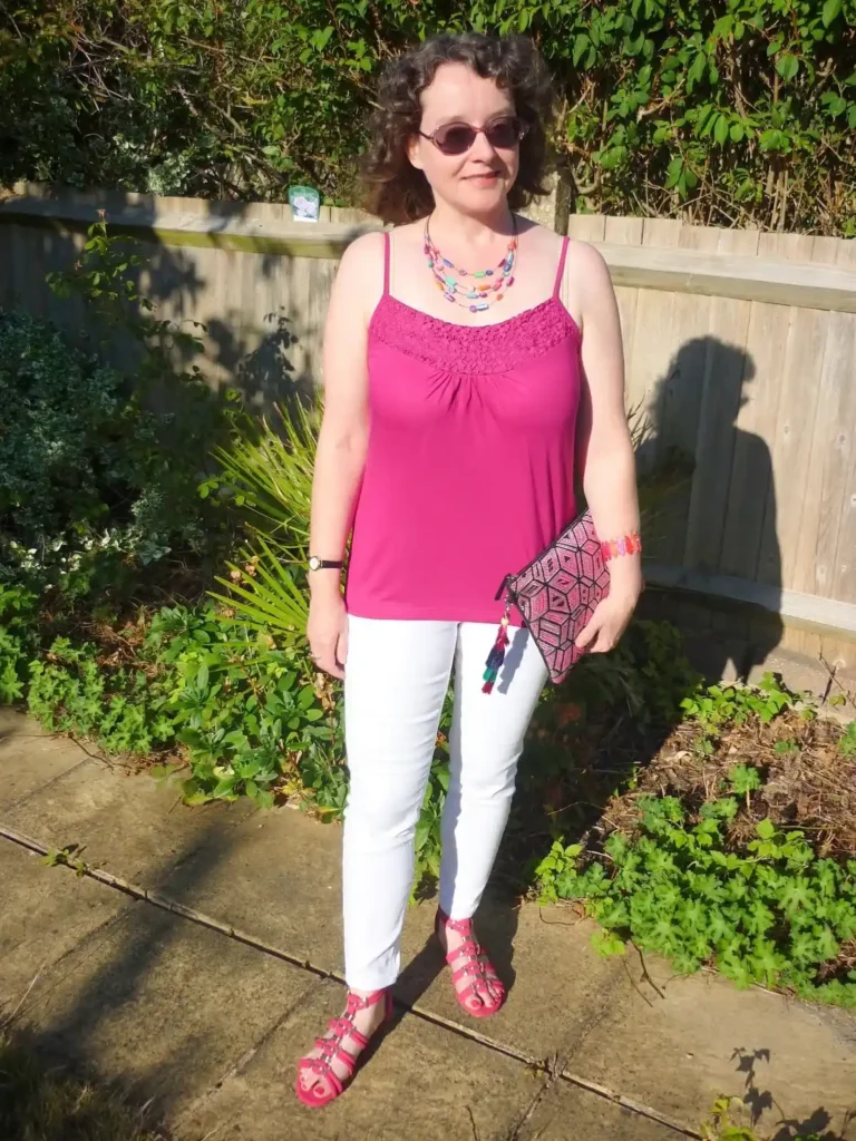 Woman with short curly dark hair and pink-tinted oval sunglasses wearing white slim-fit ankle-crop skinny jeans, a bright fuchsia magenta spaghetti-strap camisole top with a lace crochet yoke at the chest, fuchsia studded gladiator flat sandals, carrying a pink and purple printed fabric clutch pouch, with a multi-strand colorful beaded necklace and a pink beaded bracelet, standing outdoors in a garden setting with a wooden fence and green hedgerow in the background.