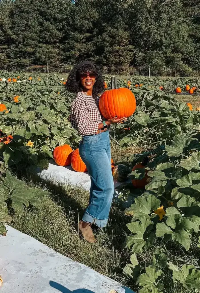 Petite woman in a brown and cream gingham blouse tucked into high-waisted medium-blue wide-leg jeans with a cuffed hem, wearing tan ankle boots and amber-tinted tortoise sunglasses, smiling while holding a large orange pumpkin in a lush green pumpkin vine field with trees in the background.