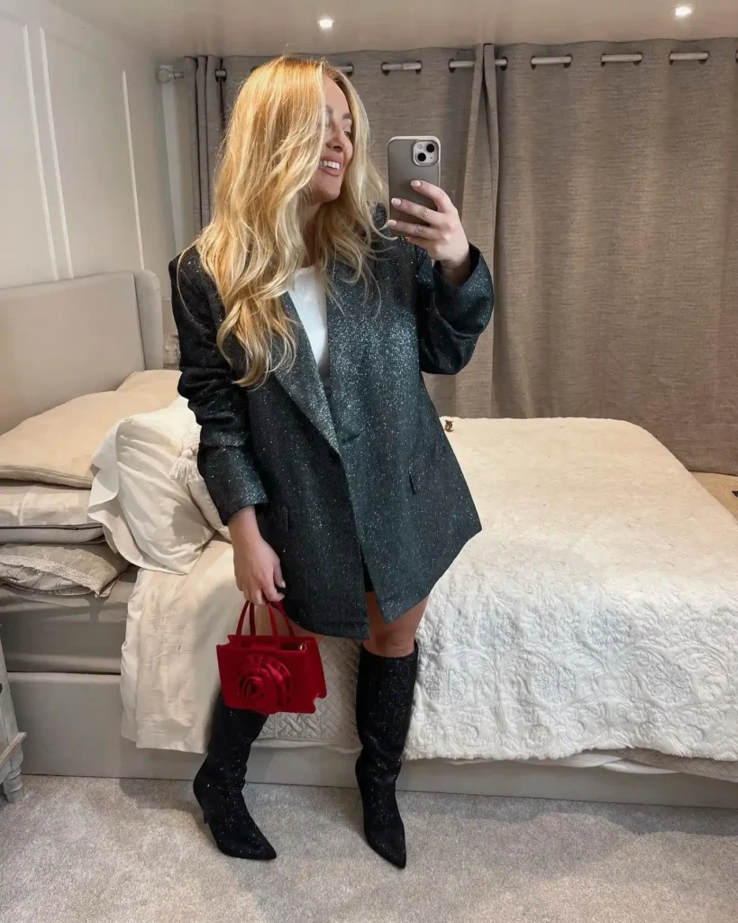 Petite woman in a dark green black glitter oversized blazer worn as a mini dress over a white top, paired with matching black sparkle glitter knee-high heeled boots and carrying a small structured red velvet mini bag with a rose appliqué detail, taking a mirror selfie in a softly lit bedroom.