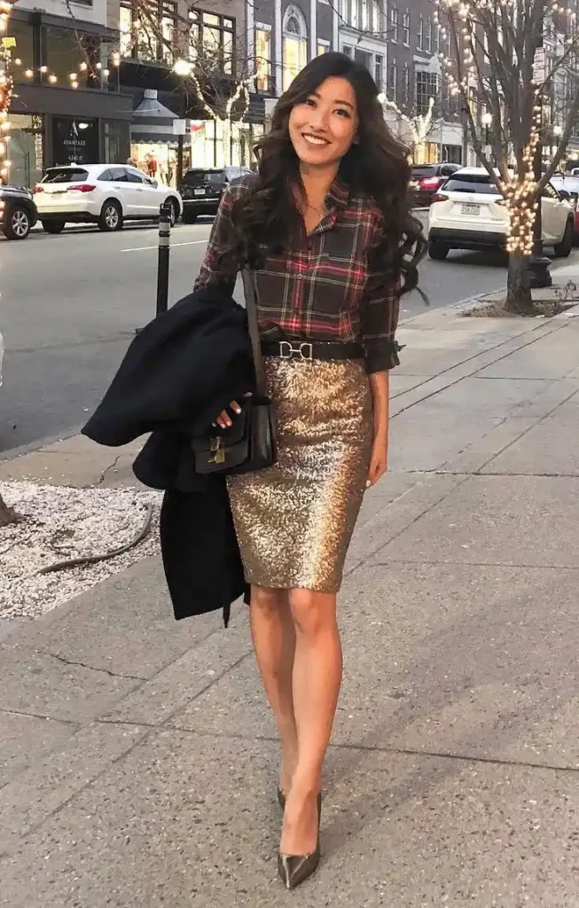 Petite woman in a dark plaid flannel shirt tucked into a gold sequin pencil skirt with a decorative belt buckle at the waist, wearing taupe metallic pointed-toe stiletto heels and carrying a small black structured crossbody bag with gold hardware and a navy coat draped over one arm, standing on a city sidewalk lined with warm fairy lights on bare winter trees.