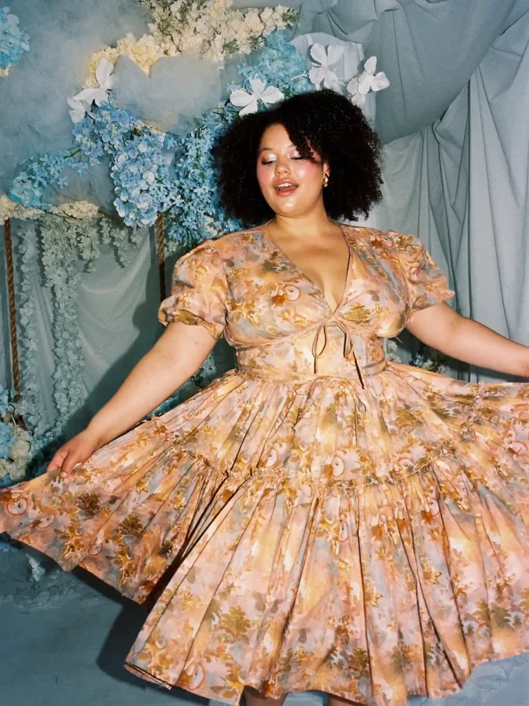 Petite plus-size woman with voluminous natural curly dark hair and small gold stud earrings, smiling with eyes slightly downcast while twirling a warm golden baroque scroll floral print short puff-sleeve tiered mini dress with a deep V-neckline and front tie detail at the bust, standing in front of a styled blue hydrangea and white flower photo backdrop with draped grey fabric.