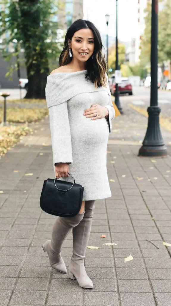 Petite pregnant Asian woman with long dark wavy highlighted hair in a light grey off-the-shoulder ribbed sweater dress with a wide folded neckline and long sleeves, wearing grey over-the-knee suede block heel boots, holding a black structured handbag with a circular metal ring handle and wearing long white tassel drop earrings, standing on a brick city sidewalk with autumn-colored trees and a street lamp in the background.