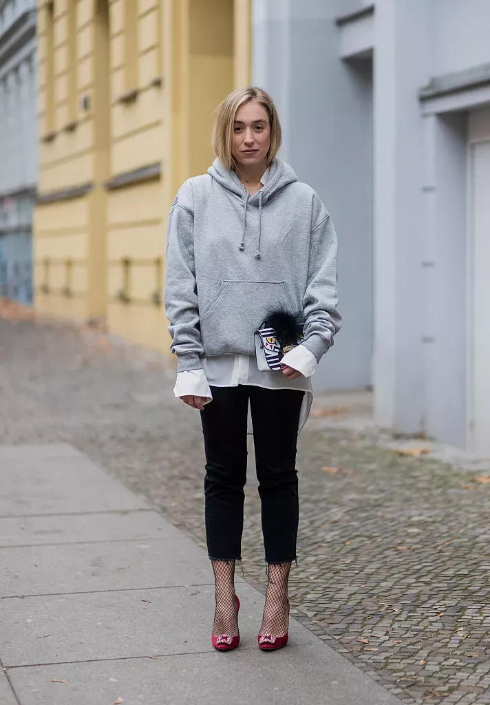 Petite woman with short blonde bob standing on a cobblestone European street, wearing black cropped straight-leg jeans with a raw frayed hem, an oversized grey pullover hoodie layered over a white button-down shirt with visible cuffs, fishnet ankle socks, and hot pink pointed-toe heels, carrying a small striped clutch with a dark feather pom detail, photographed against colourful building facades.