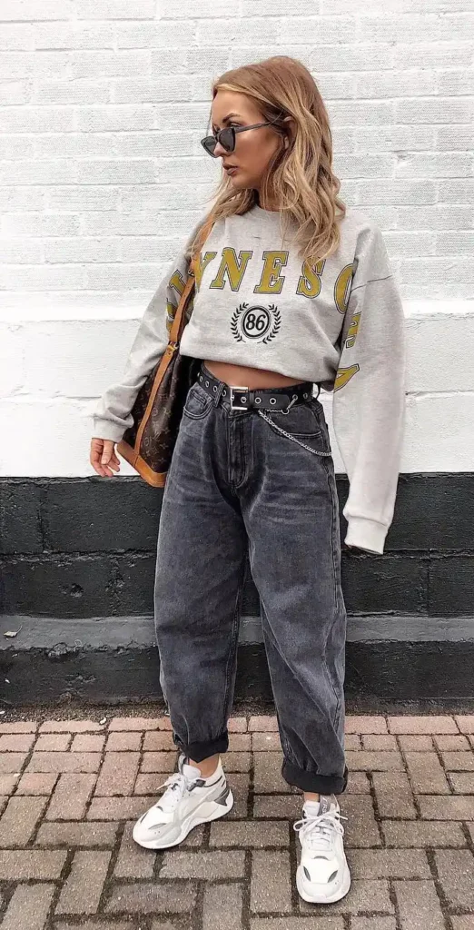 Petite woman in dark charcoal washed baggy boyfriend jeans cuffed at the ankle, wearing a cropped grey collegiate graphic sweatshirt with mustard yellow varsity lettering, a black chain-detail belt at the waist, carrying a brown monogram shoulder bag, photographed against a white painted brick wall.
