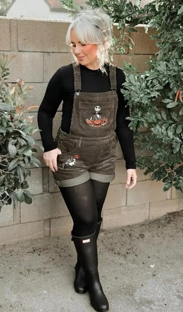 Petite woman with silver upswept hair wearing dark olive corduroy shortalls with Halloween spooky character embroidery on the bib pocket and leg panel, layered over a fitted black long-sleeve top, styled with black opaque tights and tall black rain boots, standing outdoors against a concrete wall with green foliage.