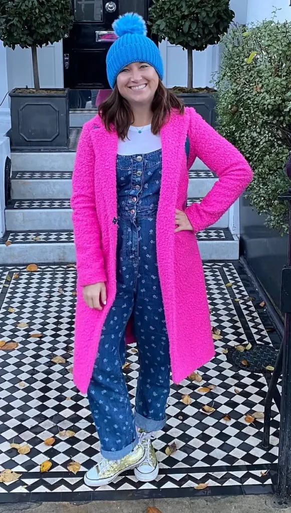 Hot Pink Teddy Coat with Floral Denim Dungarees, petite woman in open hot pink teddy coat, floral denim dungarees, blue pom beanie and gold glitter sneakers on a black and white tile path.