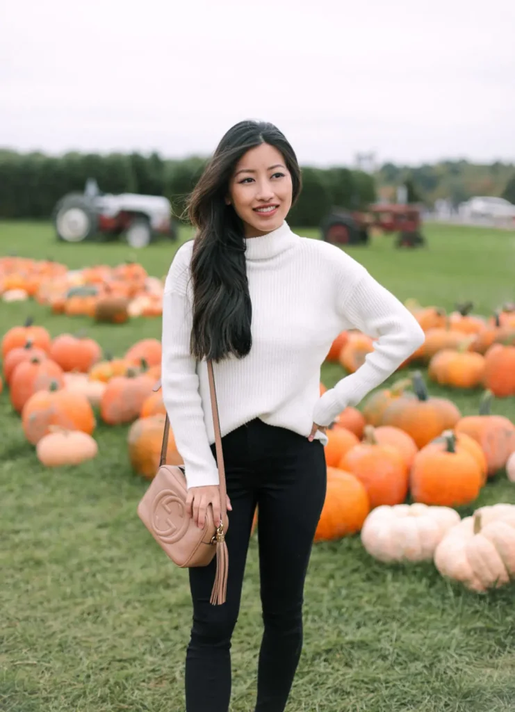Petite woman in a clean ivory ribbed turtleneck sweater with a subtle twist detail at the hem, tucked into high-waist black skinny jeans, carrying a small blush nude round crossbody bag with tassel and circular hardware, standing with one hand on her hip and a relaxed smile in a grassy pumpkin patch with orange and white pumpkins and vintage tractors visible in the background on an overcast fall day.