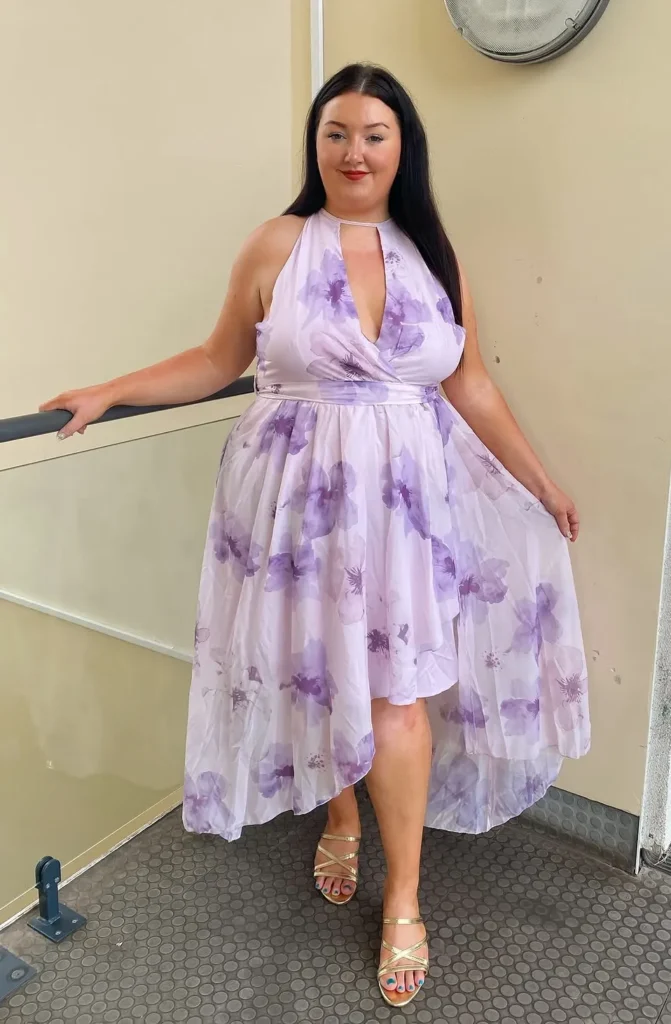 Petite plus-size woman with long straight dark hair and bold red lips, leaning lightly on a railing and smiling at the camera, wearing a sleeveless lavender and purple watercolor large-scale floral print halter midi dress with a keyhole cutout neckline, deep V wrap-style bodice, satin waistband, and a high-low chiffon skirt that is shorter at the front and trails longer at the sides and back, paired with gold cross-strap strappy heeled sandals, indoors against a neutral beige wall.
