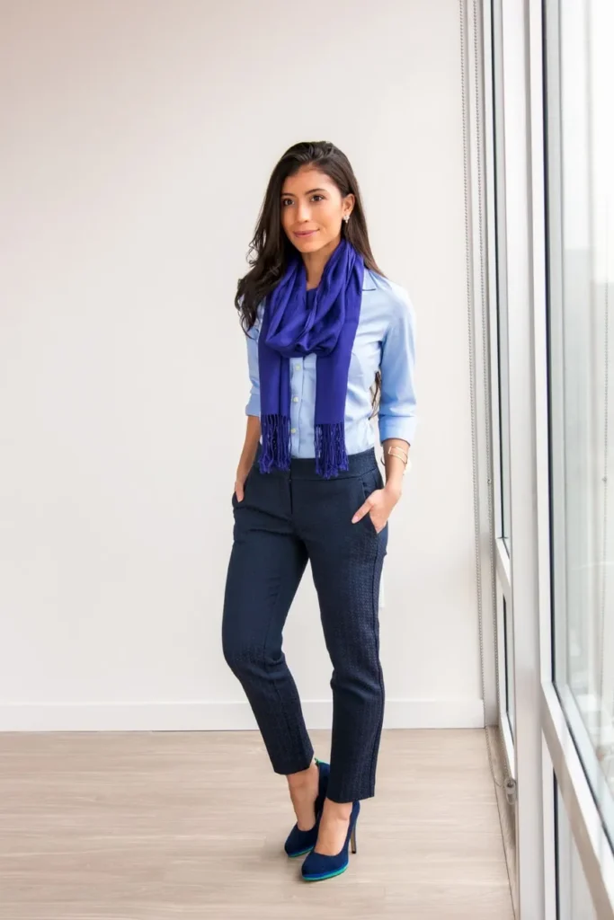 Petite woman in a light blue button-up shirt with three-quarter sleeves styled with a deep royal blue fringed scarf draped loosely around the neck, paired with slim-fit navy textured tone-on-tone jacquard cropped ankle trousers with side pockets, wearing navy suede pointed-toe pumps with a coloured sole detail, standing in a bright minimal interior next to large floor-to-ceiling windows with light wood flooring.