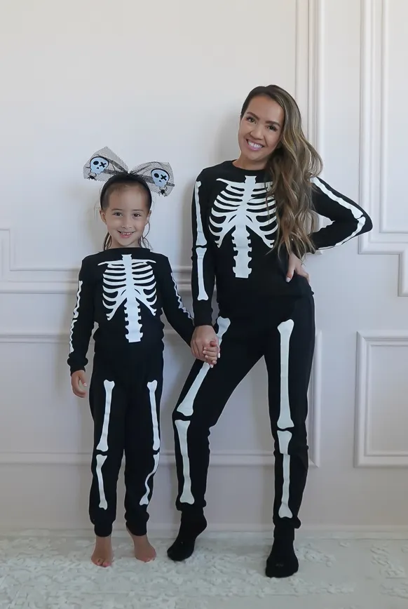 Petite woman with long wavy brunette hair wearing a black Halloween skeleton print matching set with white bone graphics across the chest, arms, and legs, paired with black sneakers, standing indoors beside a young girl in a matching child-sized skeleton print set wearing a Halloween skull tulle bow headband, both smiling against a white paneled wall.