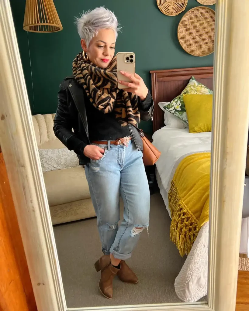 Petite woman taking a bedroom mirror selfie wearing light wash distressed boyfriend jeans with a wide ankle cuff, a cropped black leather biker jacket over a black knit sweater, a large tiger-print blanket scarf looped at the neck, a tan woven belt at the waist, a cognac leather shoulder bag, and tan ankle booties.