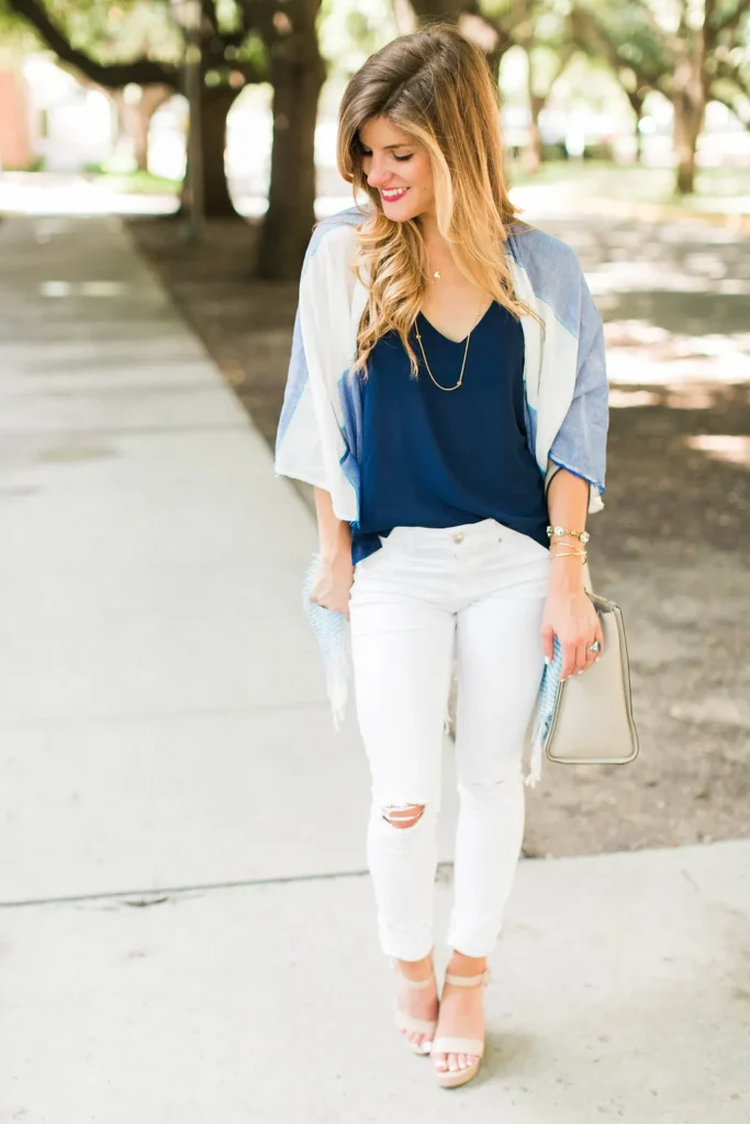 Petite woman with long wavy brunette hair wearing white ripped-knee ankle-crop skinny jeans, a loose navy blue V-neck cami top tucked lightly at the front, an open light blue and white colorblock kimono wrap with wide sleeves and a fringe hem, nude wedge sandals with an ankle strap, carrying a small structured grey taupe box clutch, with gold layered necklaces and a gold bracelet stack, walking on a sunny tree-lined sidewalk.