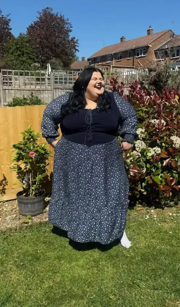 Petite plus-size woman with long dark wavy hair, laughing with eyes closed and hands tucked in her pockets, wearing a navy blue small white ditsy floral print long-sleeve maxi dress with full bishop sleeves, a dark solid navy fitted corset bodice panel with a front lace-up cord detail, and a full gathered maxi skirt in the matching print, standing on green lawn in a sunny residential backyard garden with flowering shrubs, red-leaved ornamental plants, and a wooden fence in the background, with white sneakers barely visible beneath the hem.