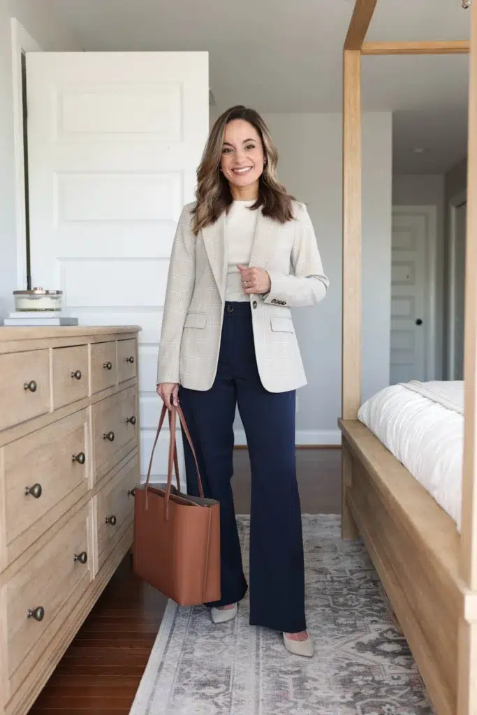 Petite woman in a fitted cream mock-neck top underneath a structured single-breasted cream and beige glen plaid check blazer with flap pockets, paired with high-waisted navy full-length flare trousers, carrying a large structured cognac tan leather tote with top handles, wearing light nude pointed-toe pumps, standing in a bright modern bedroom with a wood dresser and four-poster bed frame in the background.
