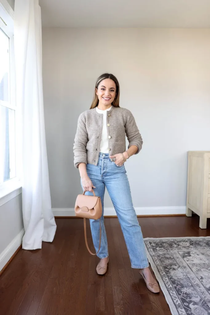 Petite woman standing in a bright home interior wearing light wash straight boyfriend jeans, a cropped oatmeal ribbed cardigan with large gold buttons over a white crew-neck top, holding a small camel tan structured top-handle bag, with nude bow detail ballet flats.