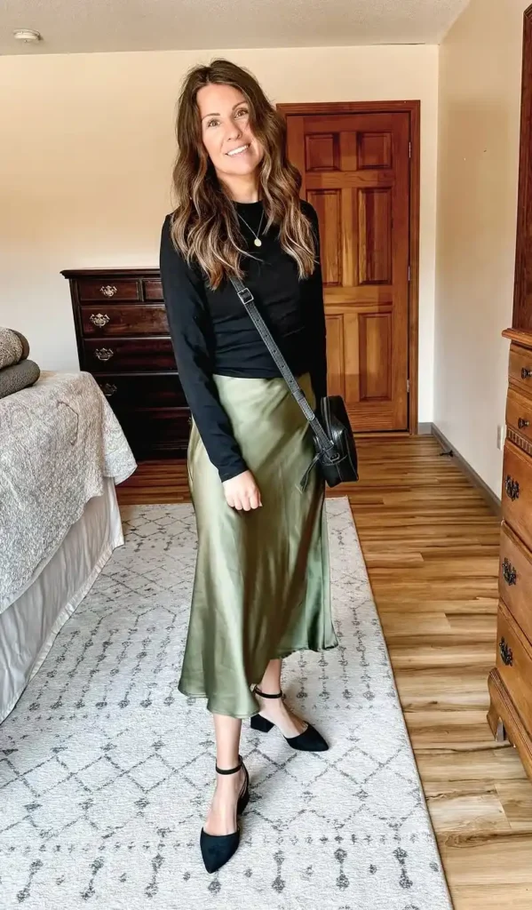Petite woman in a fitted black long-sleeve top tucked into an olive green satin bias-cut midi slip skirt, wearing black pointed-toe ankle-strap block heel pumps and a black structured crossbody bag with a delicate gold coin pendant necklace, standing in a neutral bedroom with dark wood furniture and a patterned area rug.