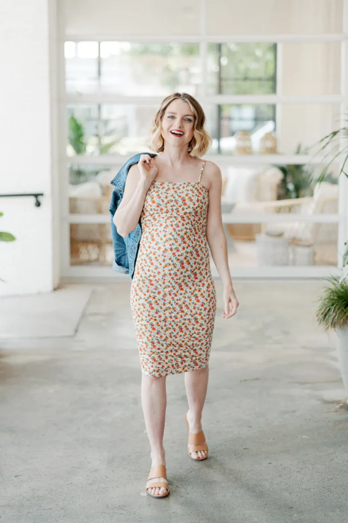 Pregnant woman with short wavy blonde hair and red lipstick in a cream and orange small-scale floral spaghetti strap fitted midi dress, carrying a light wash denim jacket over one shoulder, wearing tan block heel open-toe sandals, smiling and walking through a bright airy indoor space with large windows and neutral tones in the background.