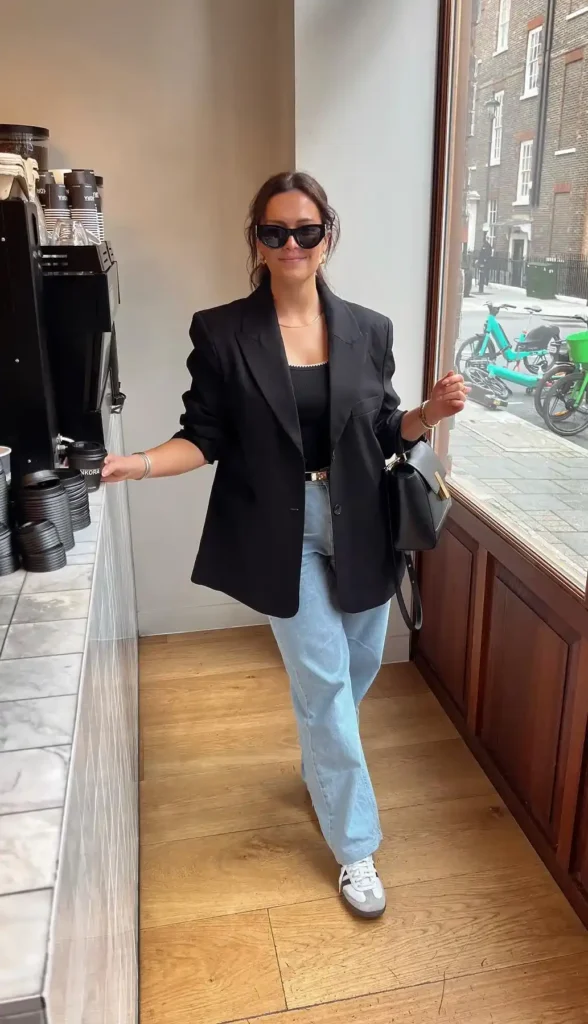 Petite woman standing in a coffee shop doorway wearing light wash relaxed straight boyfriend jeans, an oversized black blazer belted at the waist with a gold-hardware belt over a black scoop-neck cami, carrying a black structured top-handle bag, wearing white retro sneakers and black rectangular sunglasses.