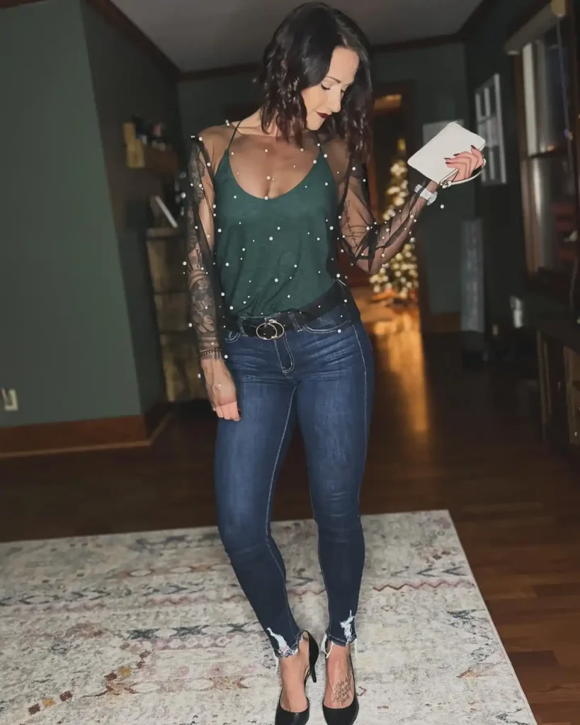 Woman in a sheer black mesh long-sleeve top with scattered pearl embellishments layered over a deep forest green V-neck cami, tucked into dark indigo skinny jeans with raw frayed ankle hems, wearing a black leather belt with a double gold ring buckle, black pointed-toe stiletto heels, and holding a small white clutch, standing in a home interior with dark green walls and a lit Christmas tree glowing warmly in the background.