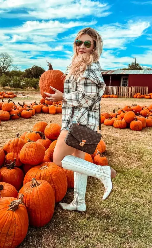 Petite woman wearing an oversized cream and grey plaid flannel shirt as a mini dress with white tall western cowboy boots and a dark brown monogram crossbody bag, wearing green mirrored aviator sunglasses, posing with one leg propped on a pumpkin in a sunny pumpkin patch with a red barn in the background.