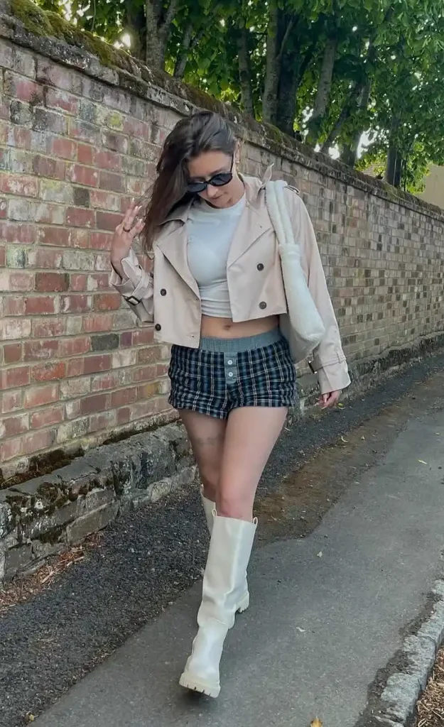 Petite woman in a navy blue and yellow tartan plaid button-front micro mini skirt with a grey ribbed waistband, layered under a cropped double-breasted pale blush trench jacket, worn over a white crop top, paired with white chunky mid-calf lug sole boots and a white fluffy sherpa shoulder bag, walking along a brick-walled street with trees overhead.
