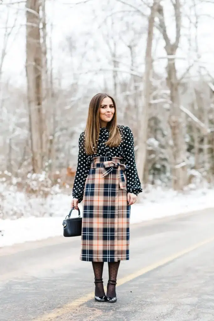Polka Dot Blouse with Plaid Bow Midi Skirt, petite woman in black polka dot blouse and high-waisted navy plaid midi skirt with self-tie bow, dotted sheer tights and black heels on a snowy road.